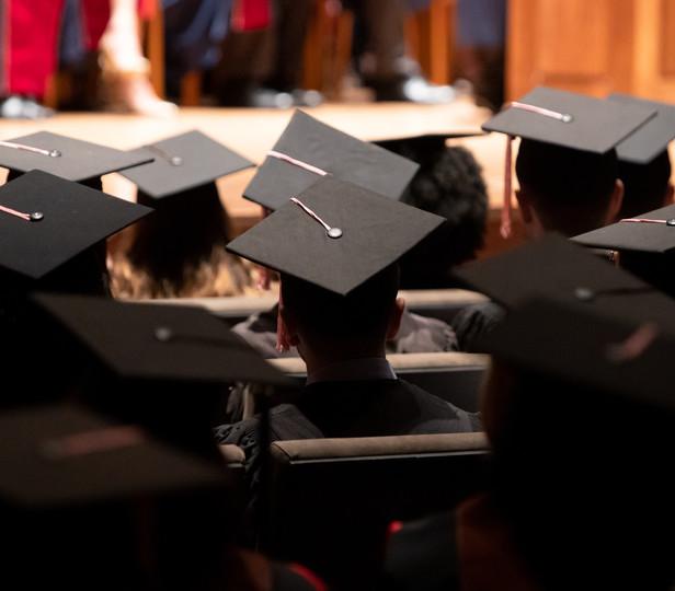 A group of graduates during a commencement ceremony. They are wearing traditional black caps and gowns. 背景是爱丽丝塔利大厅，一排排的座位上坐满了毕业生. 这张照片的角度说明是从大厅后面拍的, 重点是毕业生的后脑勺和学位帽, with one in the foreground serving as a focal point.